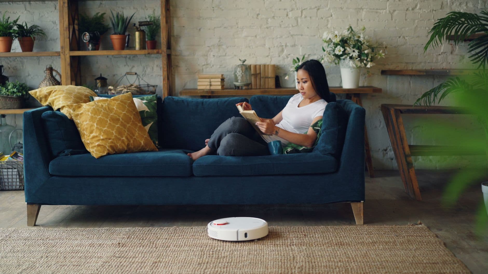 A robot vacuum cleaning a living room floor while someone reads on the sofa