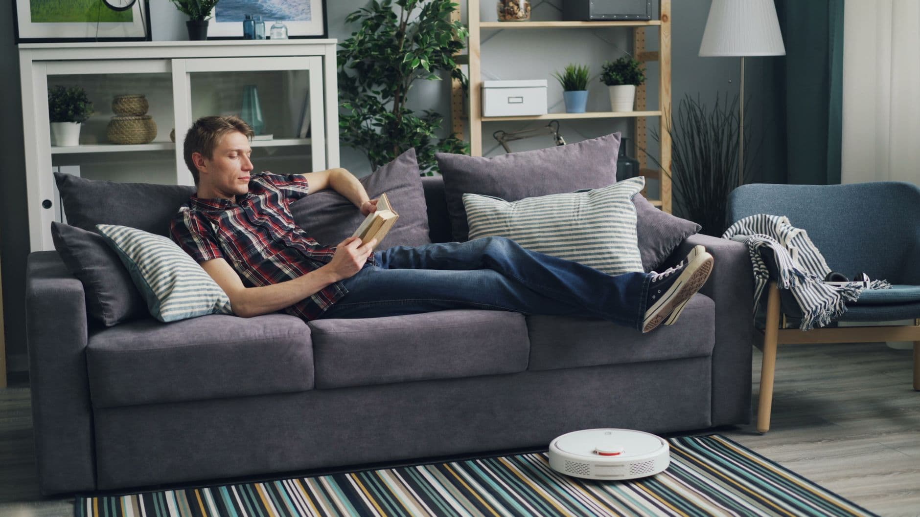 A robot vacuum cleaning a living room floor while someone reads on a sofa