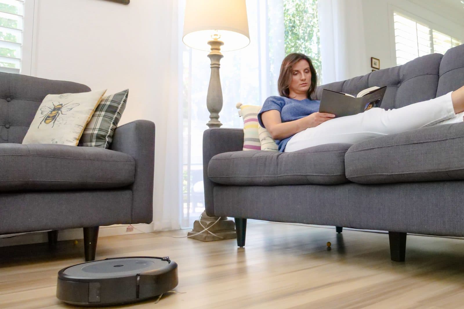 A robot vacuum cleaning a hardwood floor while someone reads on a sofa
