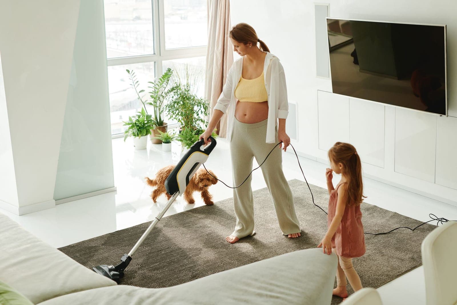 A person vacuuming a carpet with an upright vacuum while a child and dog play nearby