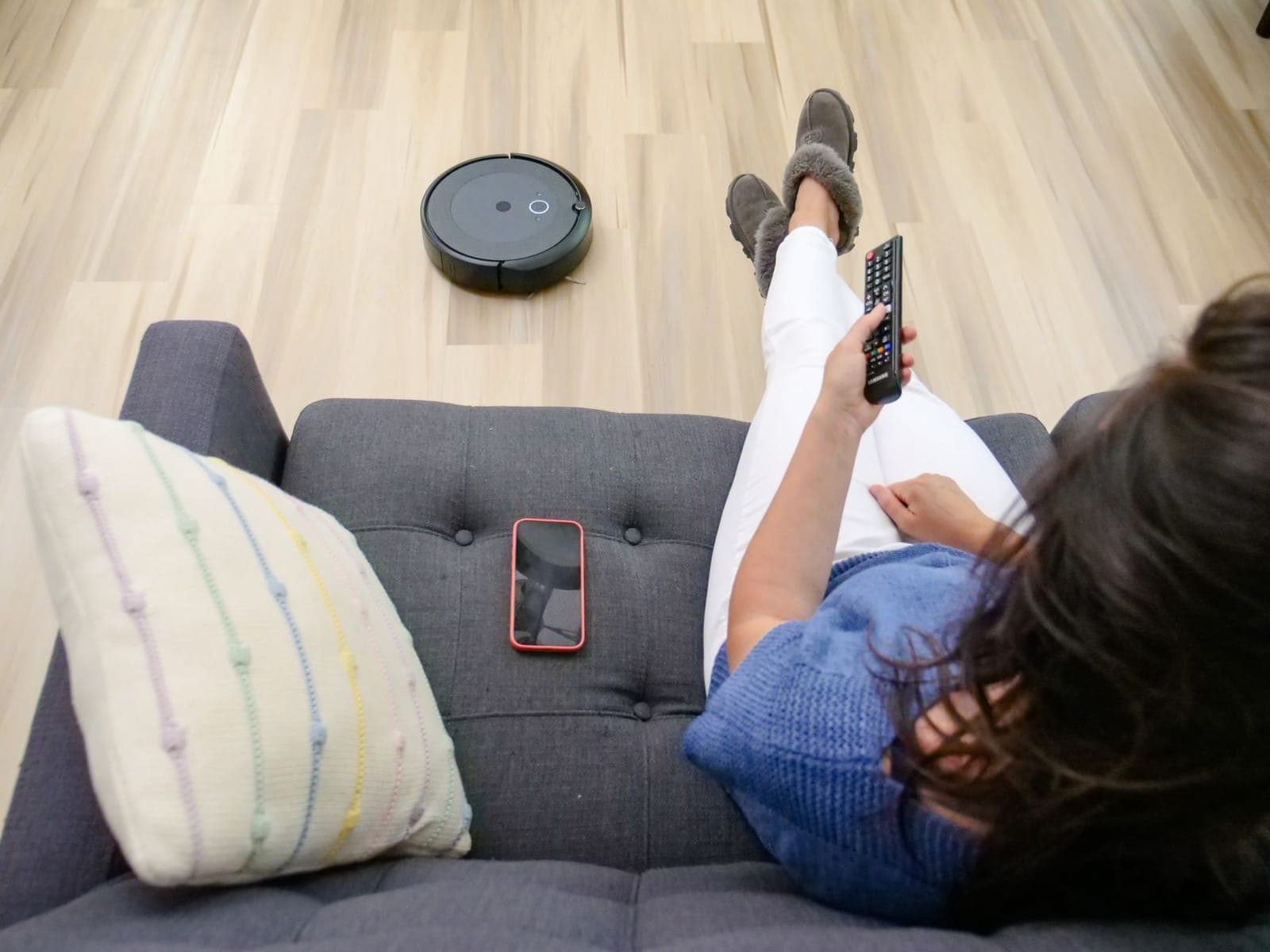 A person on a sofa using a remote while a robot vacuum cleans the floor nearby